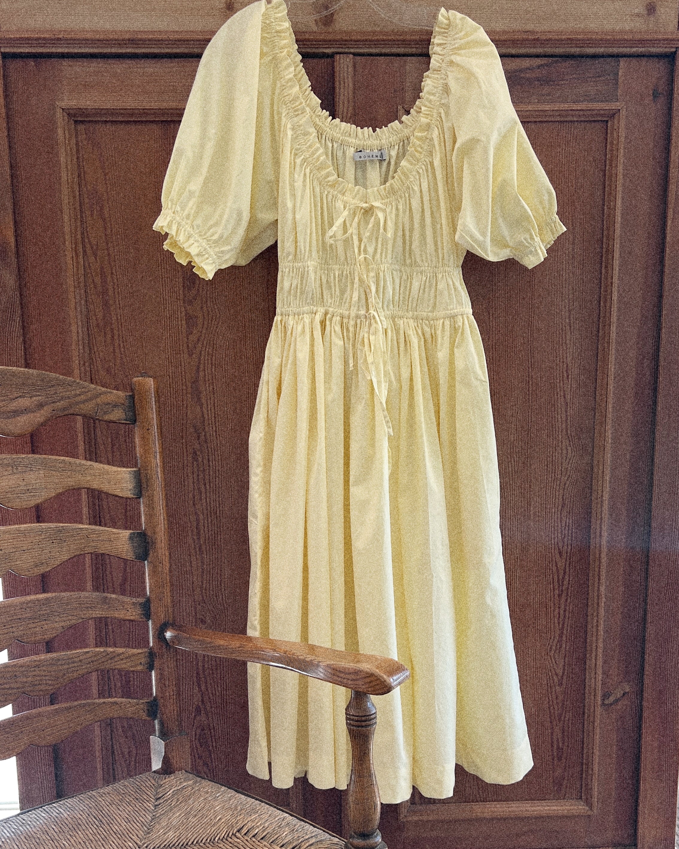 Elle boheme butter yellow colour romantic silhouette vintage inspired spring dress made of pure cotton poplin featuring a delicate scoop neckline ruffled sleeves hanging on a wooden chair against a cottage wooden panel background