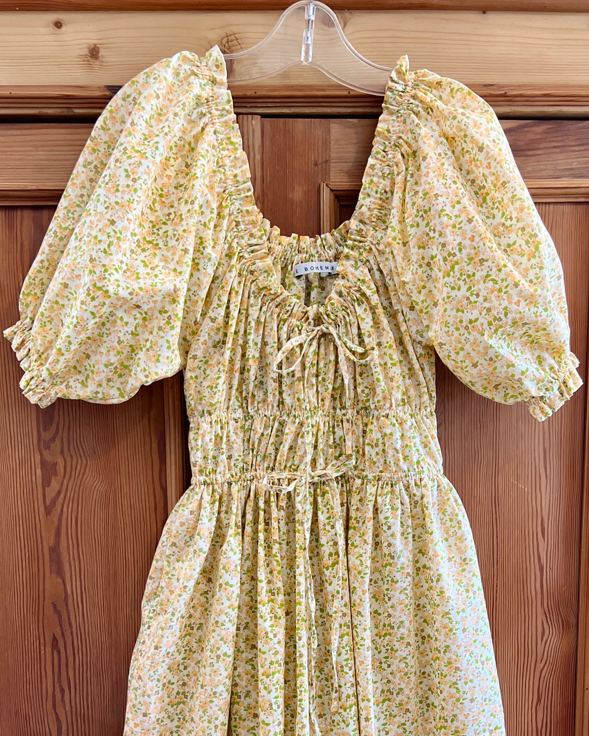 Elle boheme soft spring tones with wildflowers fabric pattern romantic dress with puff sleeves and adjustable drawstring on a hanger against a vintage pine wooden backgrounding. 