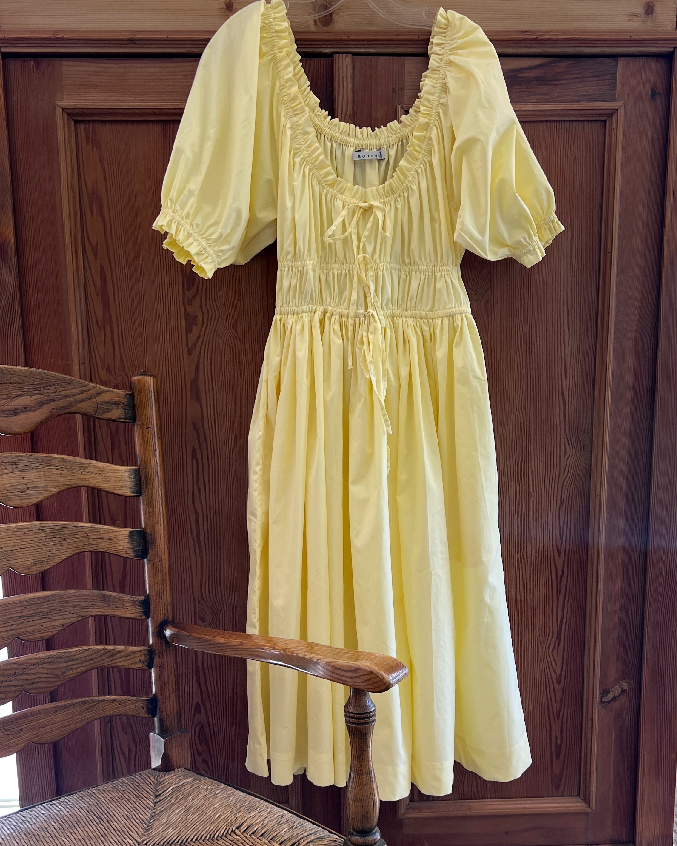 Elle boheme butter yellow colour vintage inspired spring summer pure cotton midi dress with a delicate scoop neckline ruffled sleeves and adjustable waist band hanging on a cottage wooden door.