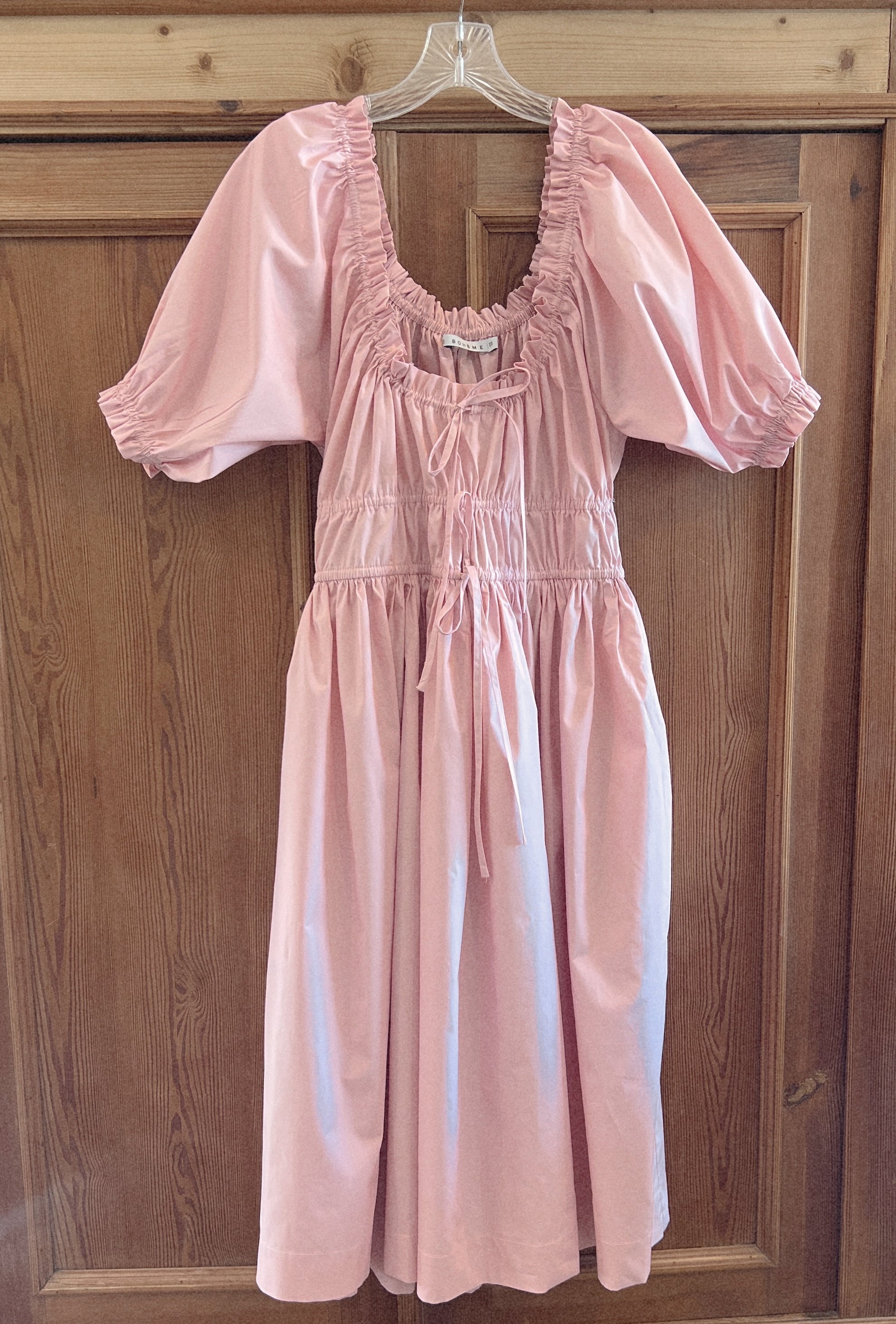 elle boheme pastel pink poplin fabric midi dress with puff sleeves and adjustable drawstring waist hanging on a english pine wooden door