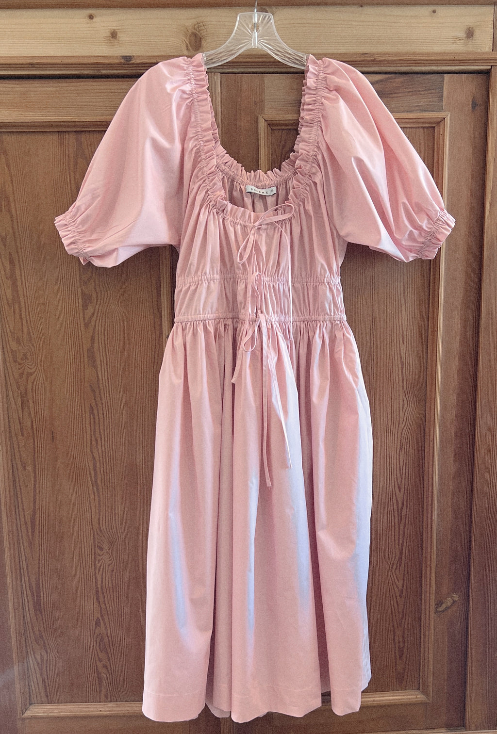 elle boheme pastel pink poplin fabric midi dress with puff sleeves and adjustable drawstring waist hanging on a english pine wooden door